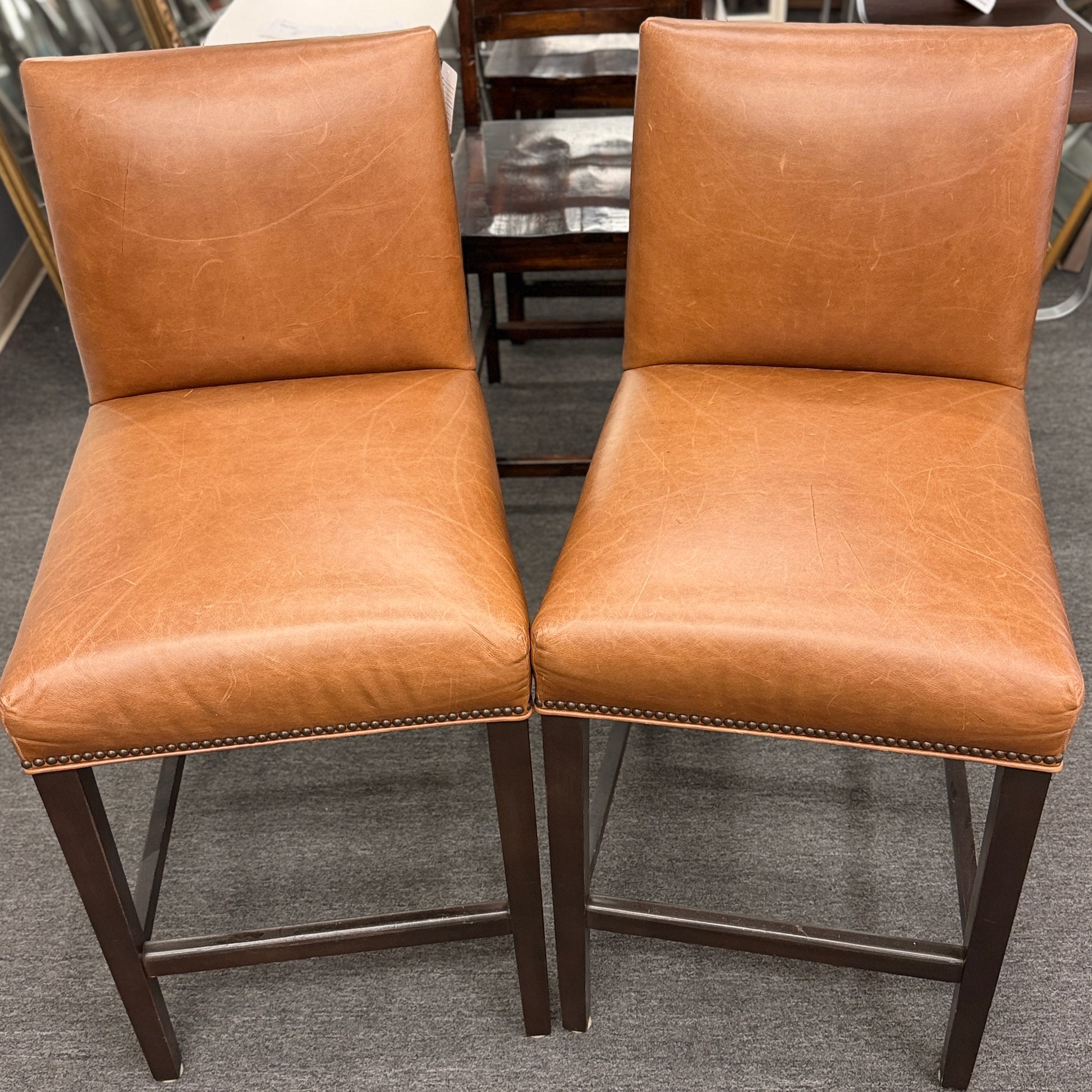 A set of two brown leather barstools featuring nailhead trim details and dark wood legs in good condition.