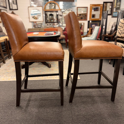 A set of two brown leather barstools featuring nailhead trim details and dark wood legs in good condition.