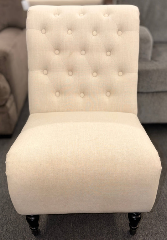 Button tufted slipper chair upholstered in cream textured fabric with an armless design, turned wooden front legs, and decorative casters.