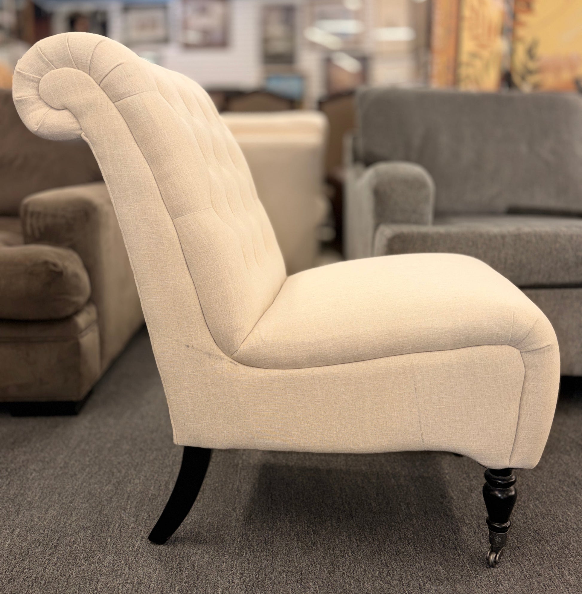 Button tufted slipper chair upholstered in cream textured fabric with an armless design, turned wooden front legs, and decorative casters.