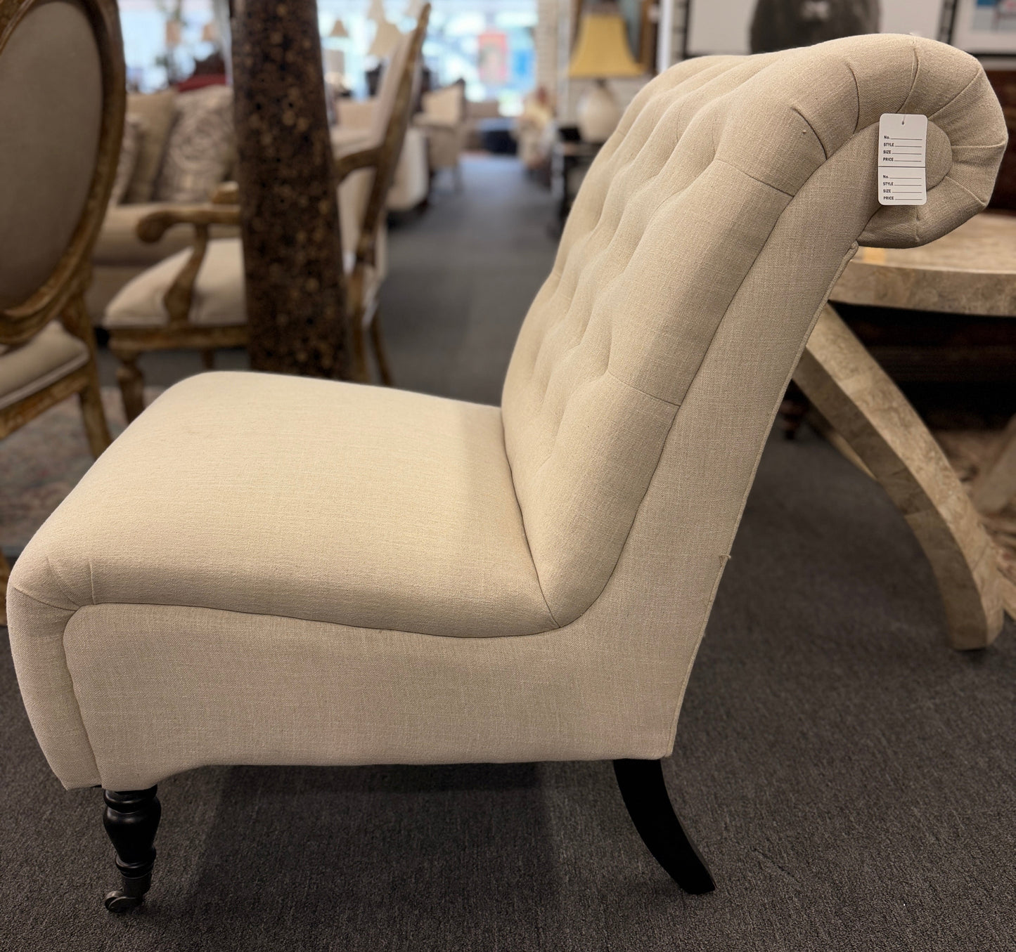 Button tufted slipper chair upholstered in cream textured fabric with an armless design, turned wooden front legs, and decorative casters.
