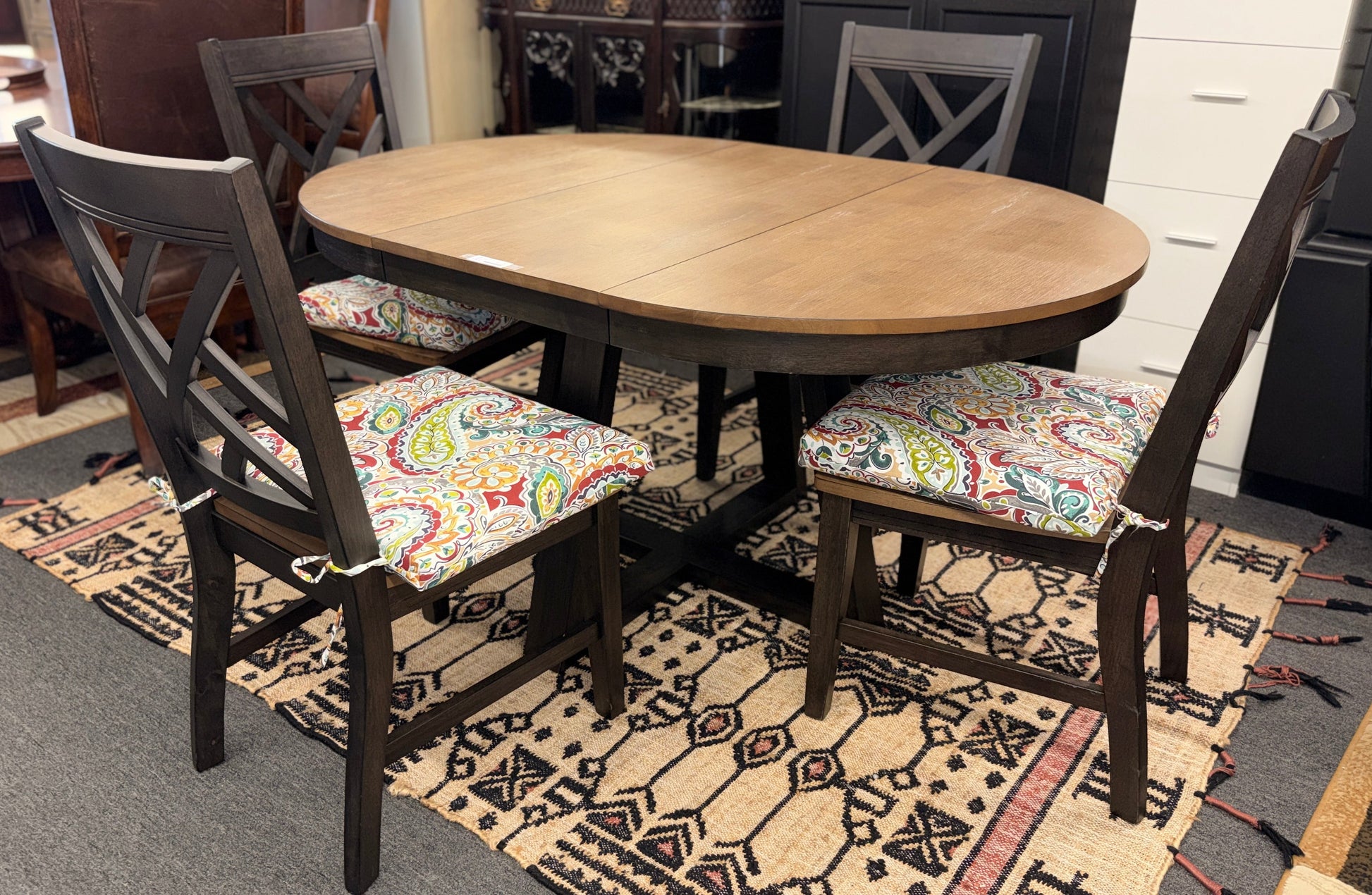 A casual oval dining table set featuring a mid-tone wood top, a dark base with an extendable leaf, and four matching X-back chairs with vibrant paisley-patterned tie-on cushions.