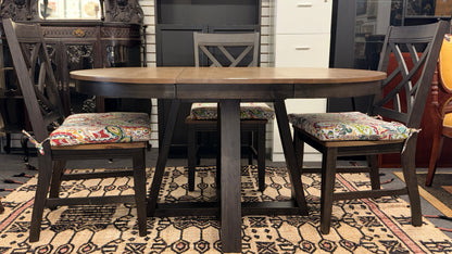 A casual oval dining table set featuring a mid-tone wood top, a dark base with an extendable leaf, and four matching X-back chairs with vibrant paisley-patterned tie-on cushions.