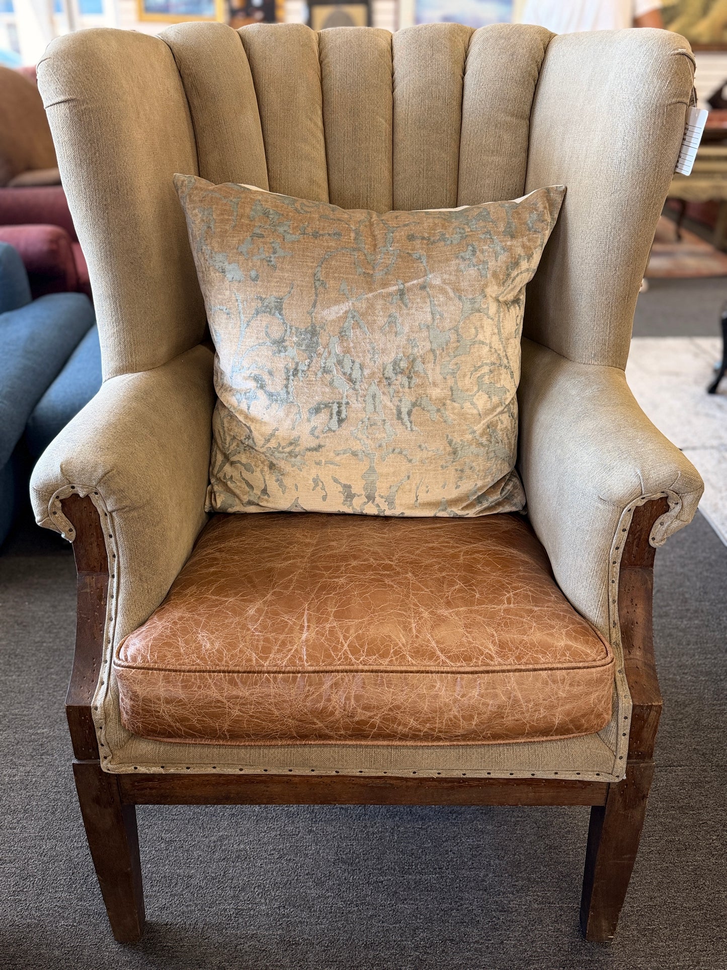 A classic wingback armchair featuring a beige channel-tufted fabric back, distressed brown leather seat cushion, dark wooden legs, and decorative nailhead trim.