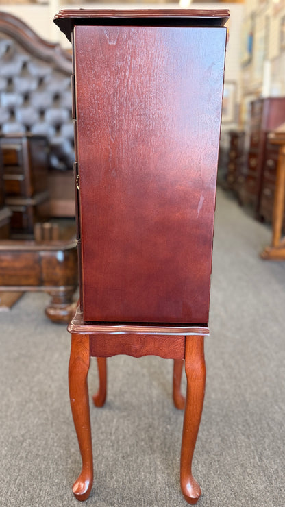 A tall cherry-finished wooden jewelry cabinet on cabriole legs featuring pull-out drawers, swinging side doors, and a flip-top vanity mirror.