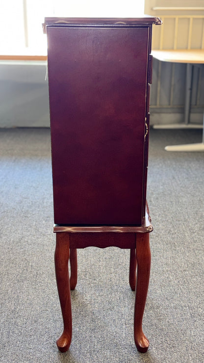 A tall cherry-finished wooden jewelry cabinet on cabriole legs featuring pull-out drawers, swinging side doors, and a flip-top vanity mirror.