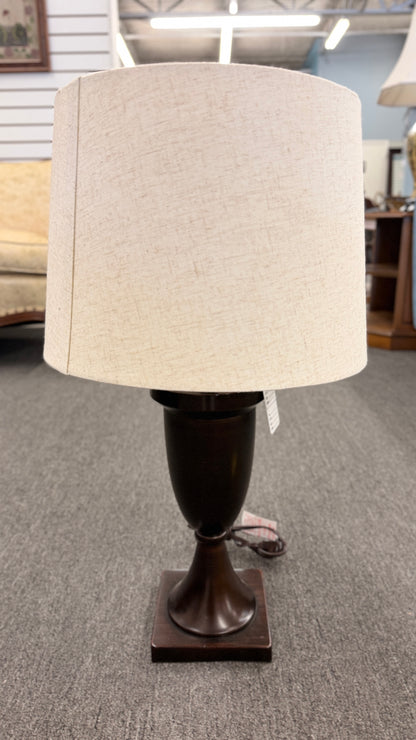 A classic table lamp featuring a dark urn-shaped iron base on a square pedestal topped with a light beige textured drum shade.