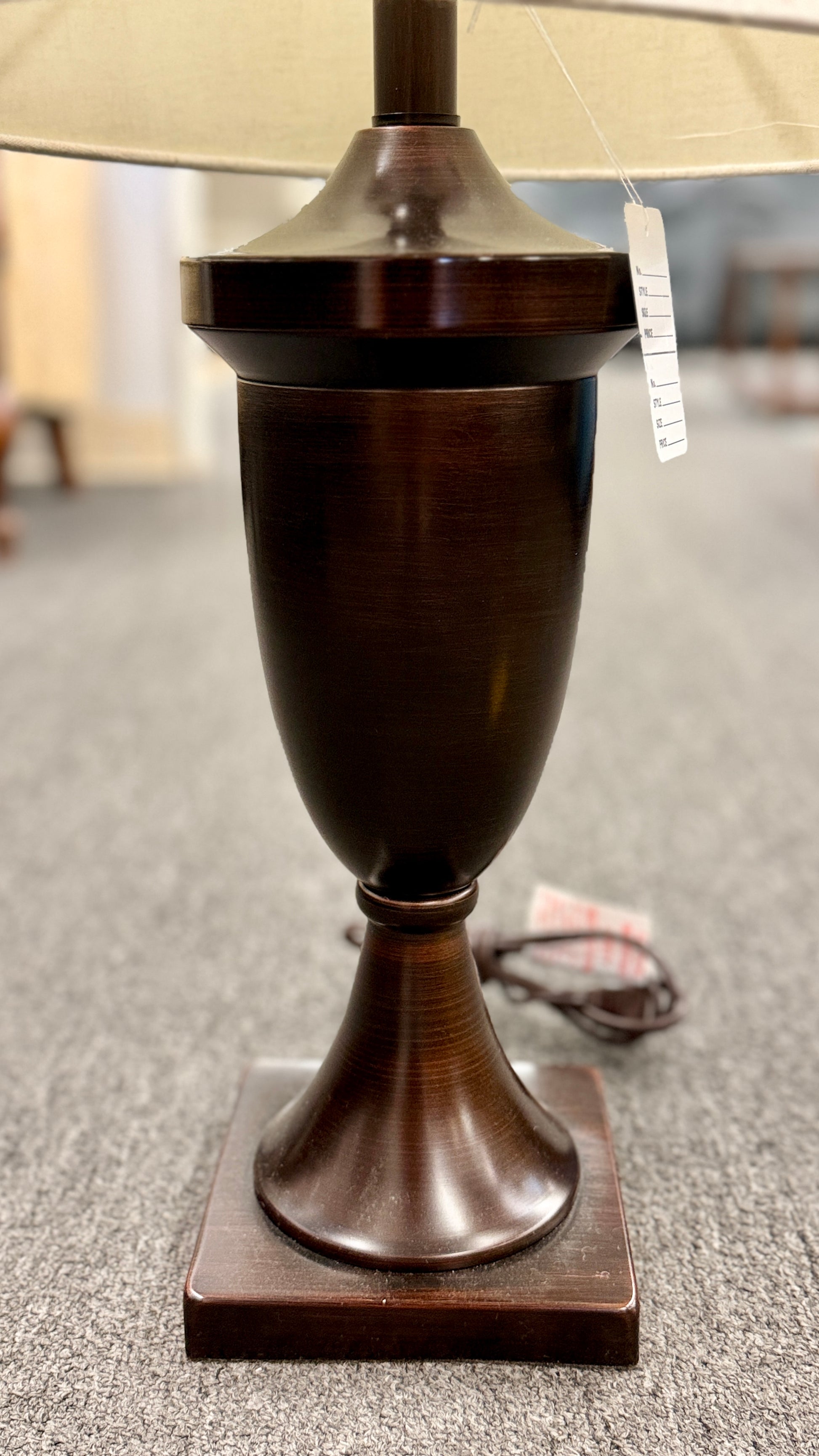 A classic table lamp featuring a dark urn-shaped iron base on a square pedestal topped with a light beige textured drum shade.
