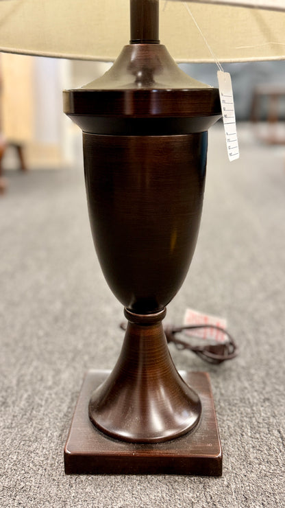 A classic table lamp featuring a dark urn-shaped iron base on a square pedestal topped with a light beige textured drum shade.