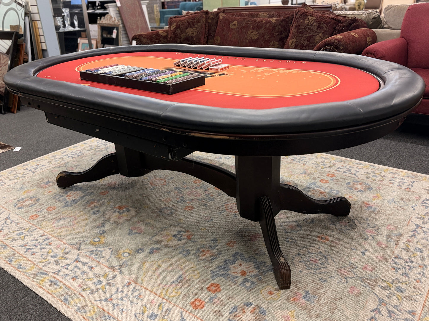 An oval-shaped Texas Hold'em poker table featuring a bright orange and red branded felt surface, black padded armrest ring, wood base, poker chips, and playing cards.