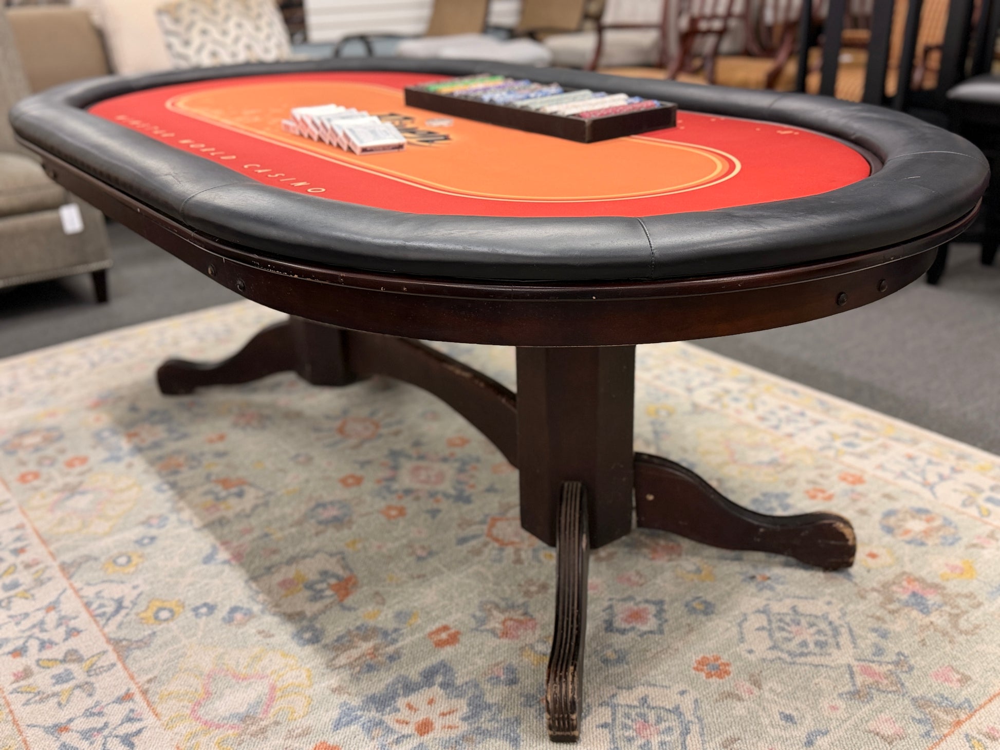 An oval-shaped Texas Hold'em poker table featuring a bright orange and red branded felt surface, black padded armrest ring, wood base, poker chips, and playing cards.