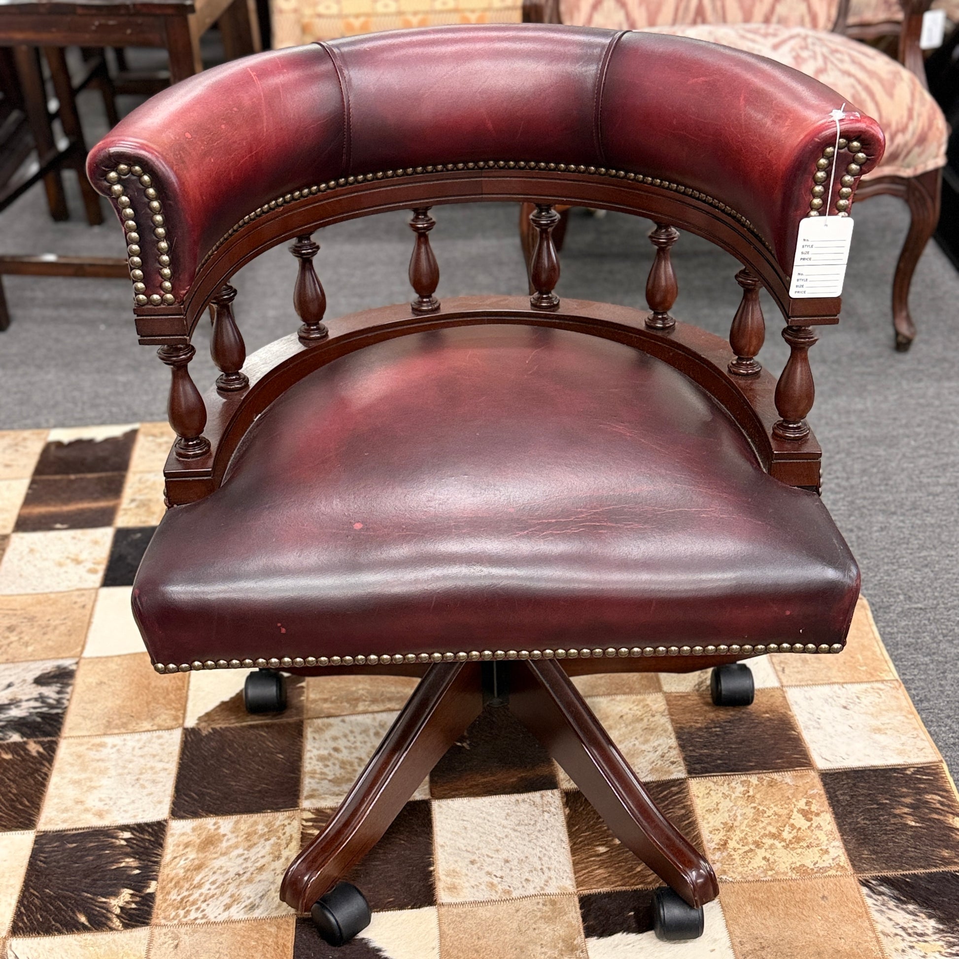 A classic traditional rolling captain's desk chair featuring a curved back, wooden spindle supports, dark leather upholstery, and brass nailhead detailing.