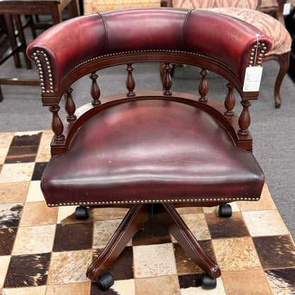 A classic traditional rolling captain's desk chair featuring a curved back, wooden spindle supports, dark leather upholstery, and brass nailhead detailing.