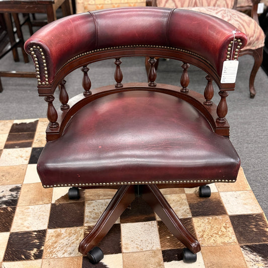 A classic traditional rolling captain's desk chair featuring a curved back, wooden spindle supports, dark leather upholstery, and brass nailhead detailing.