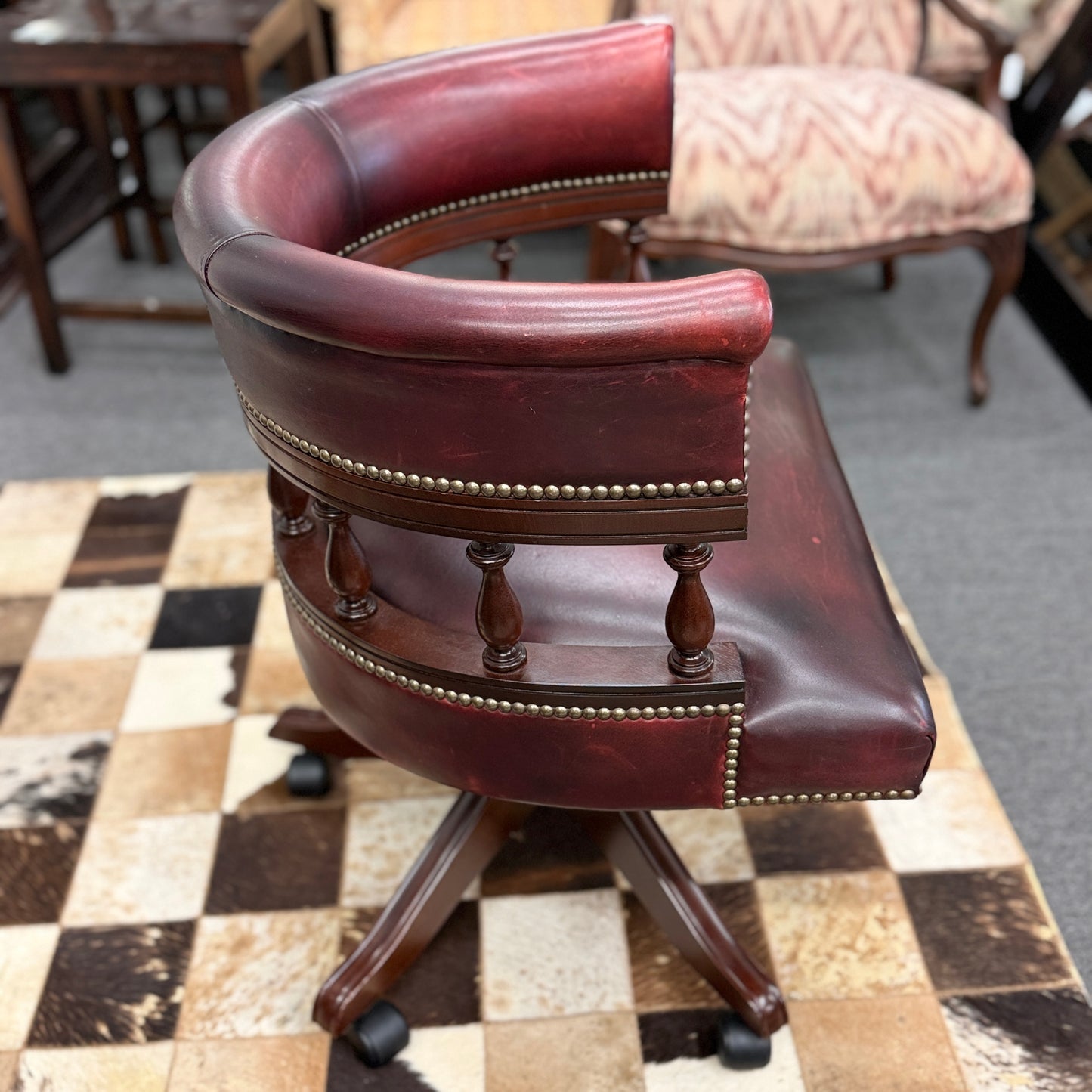 A classic traditional rolling captain's desk chair featuring a curved back, wooden spindle supports, dark leather upholstery, and brass nailhead detailing.