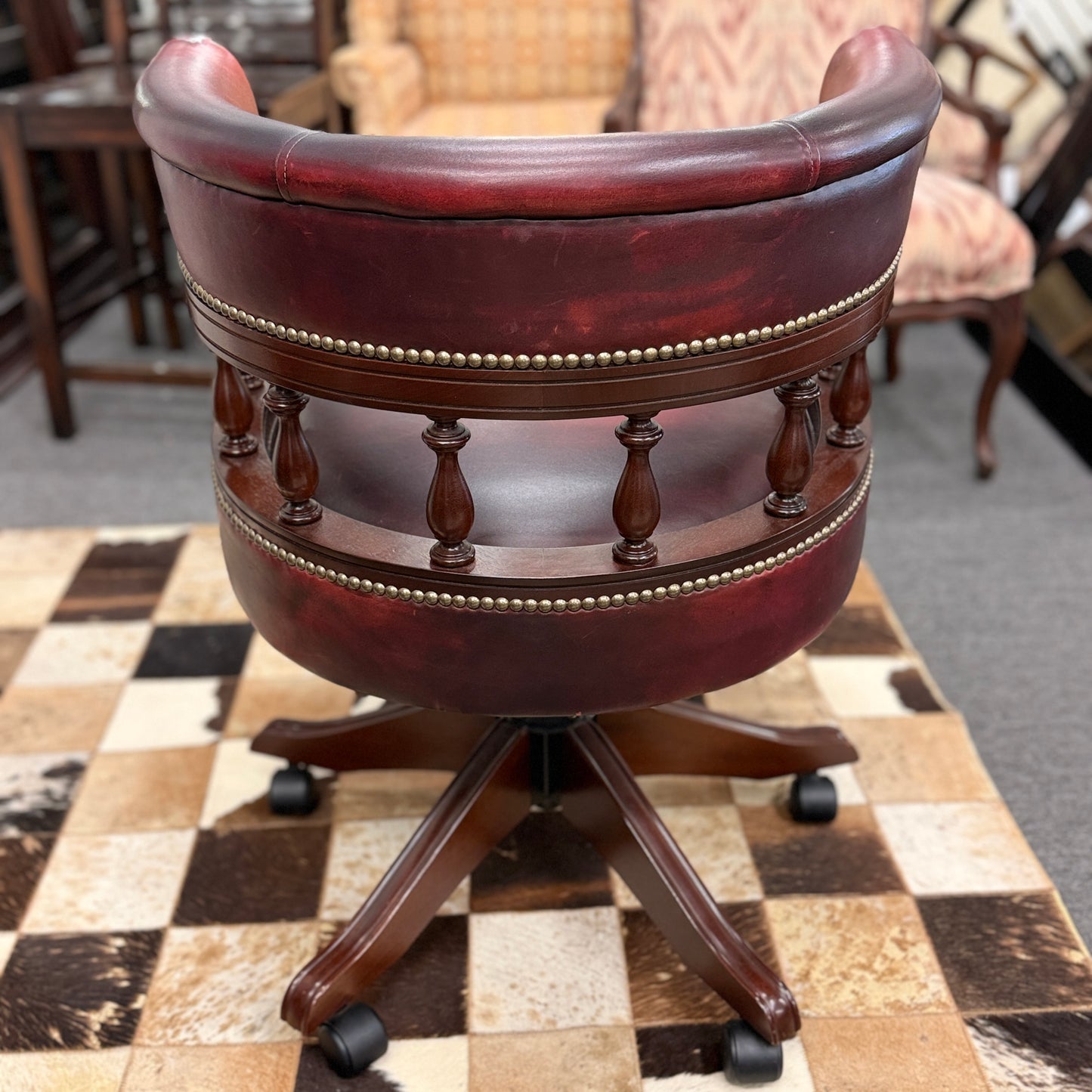 A classic traditional rolling captain's desk chair featuring a curved back, wooden spindle supports, dark leather upholstery, and brass nailhead detailing.