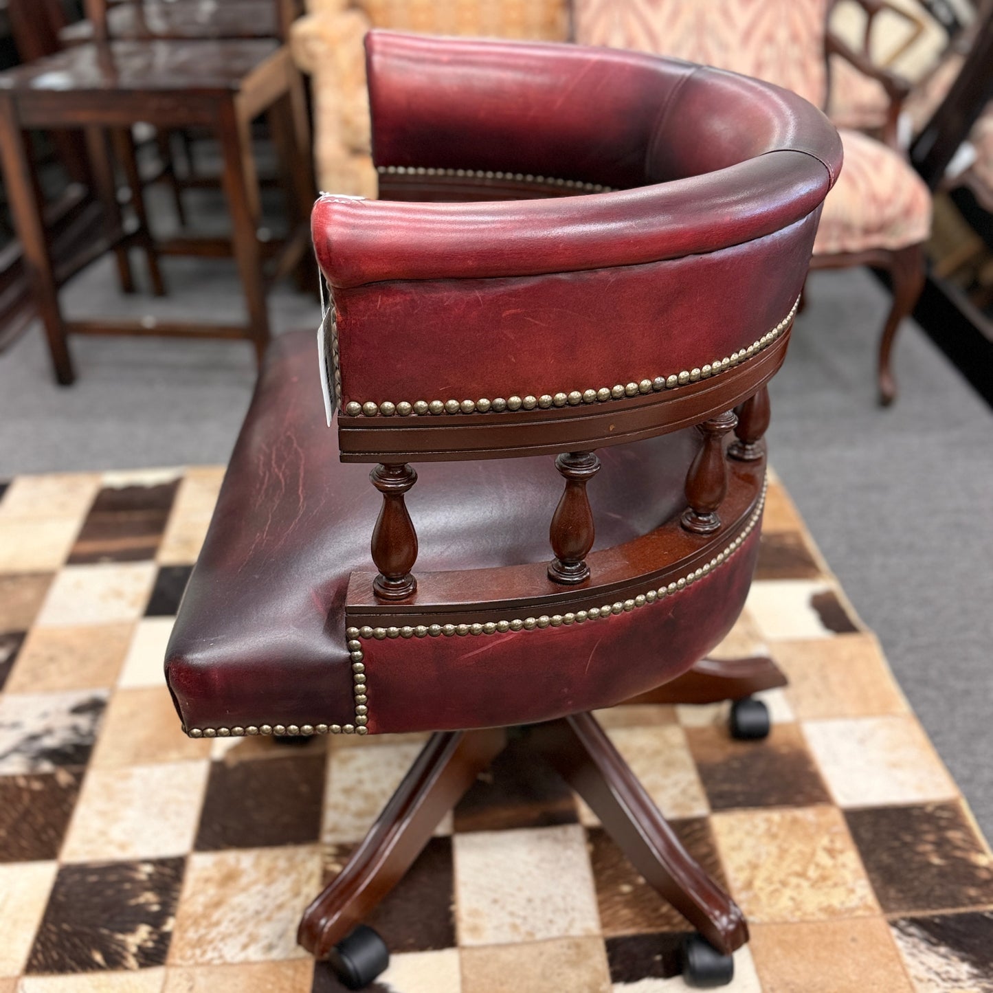 A classic traditional rolling captain's desk chair featuring a curved back, wooden spindle supports, dark leather upholstery, and brass nailhead detailing.