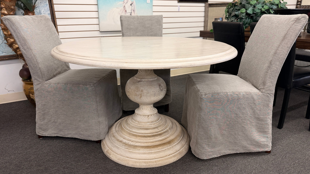 A coastal-farmhouse dining set featuring a round whitewashed pedestal table and four light gray skirted slipcover chairs in good condition.