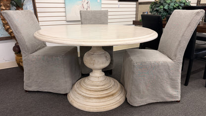 A coastal-farmhouse dining set featuring a round whitewashed pedestal table and four light gray skirted slipcover chairs in good condition.