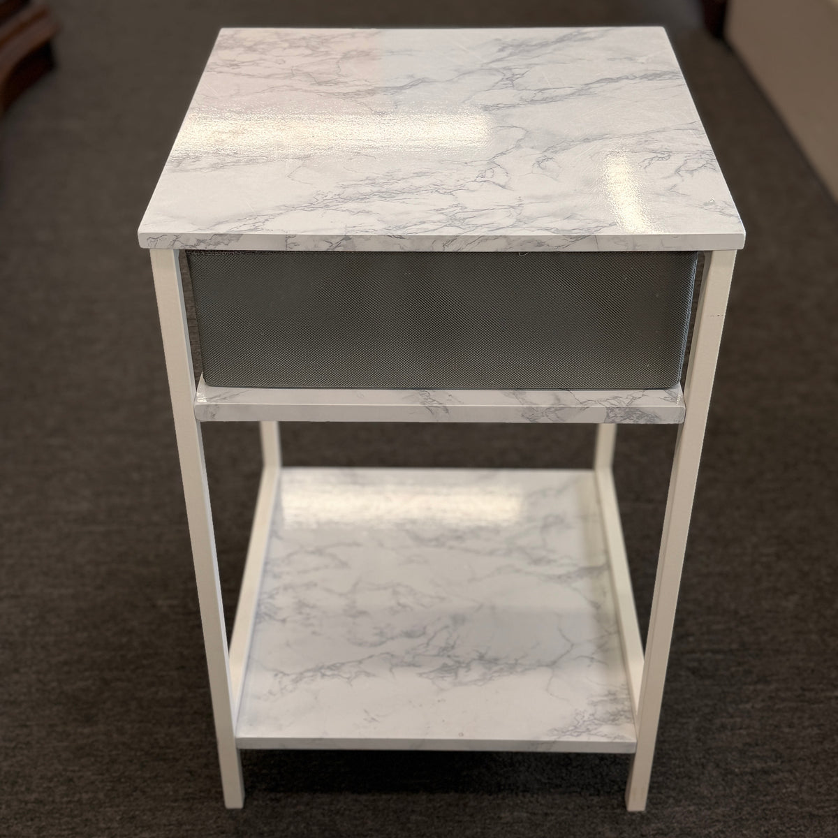 A compact nightstand featuring a white metal frame, faux marble top and bottom shelves, and a gray fabric drawer with a wooden handle in good condition.