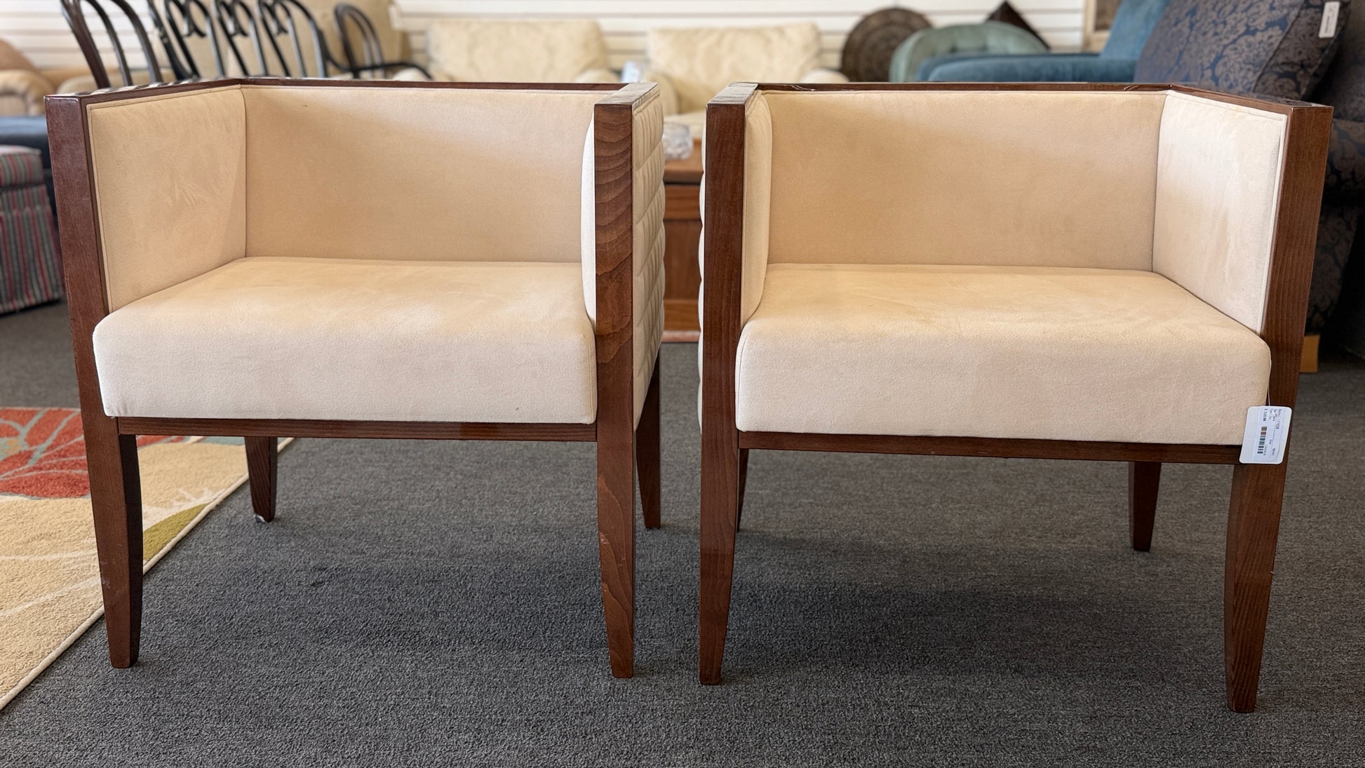 A pair of contemporary squared-off accent chairs with light beige suede-like upholstery, an exposed dark wood outer frame, and subtle quilted tufting on the side panels.