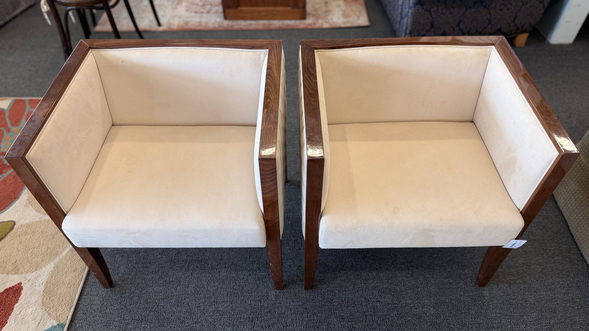 A pair of contemporary squared-off accent chairs with light beige suede-like upholstery, an exposed dark wood outer frame, and subtle quilted tufting on the side panels.