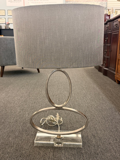 A contemporary table lamp featuring an intertwined dual-ring brushed nickel body, a clear acrylic base, and a textured gray drum shade in excellent condition.