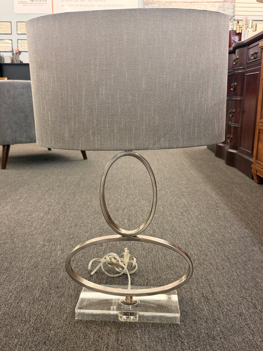 A contemporary table lamp featuring an intertwined dual-ring brushed nickel body, a clear acrylic base, and a textured gray drum shade in excellent condition.