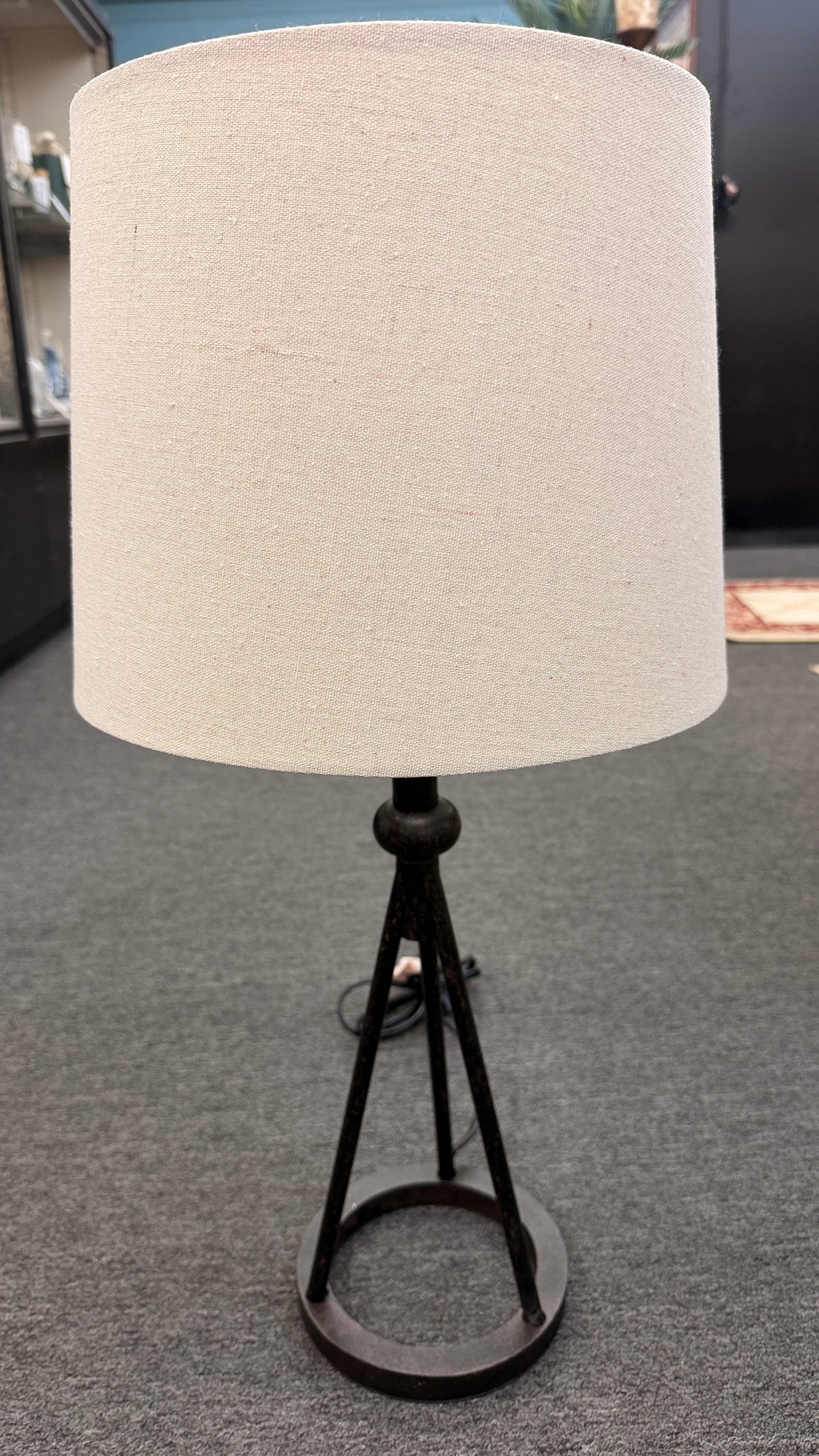 A contemporary minimalist table lamp with a dark bronze or black tripod metal base on a ring stand, topped with an oversized cylindrical off-white linen drum shade in good condition.