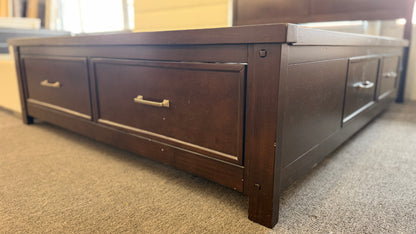 A contemporary dark-finished queen bed frame featuring a simple paneled headboard, wooden slat support, and two built-in storage drawers in the footboard.