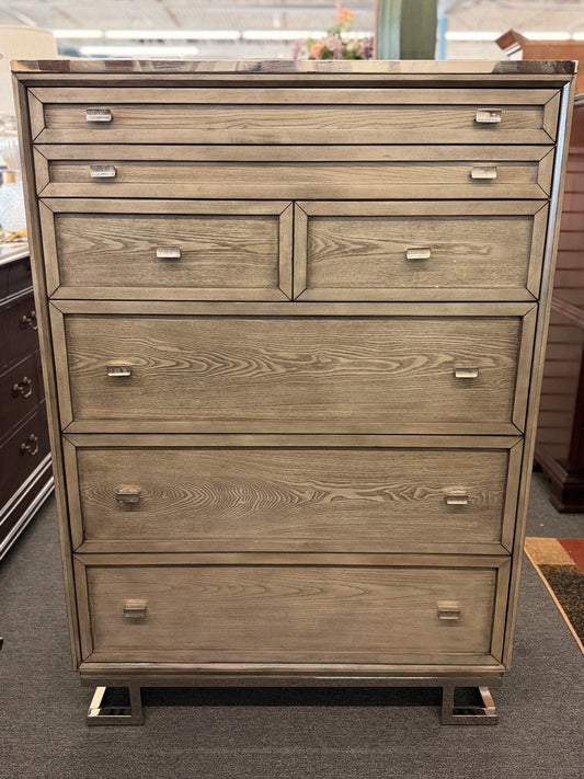 A contemporary tall 7-drawer highboy chest of drawers with a soft gray-washed wood grain finish, minimalist silver hardware, and a geometric chrome base.