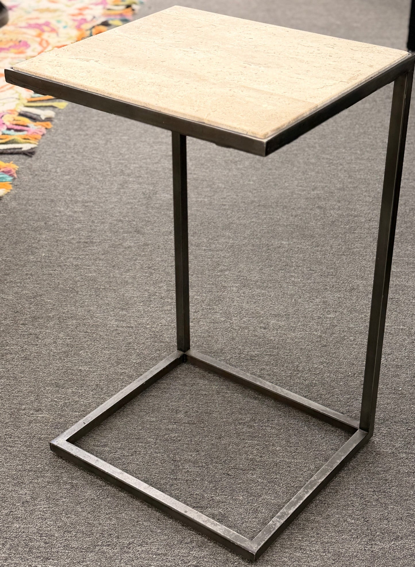 A contemporary C-shaped minimalist side table featuring a square tan stone top on a slender black metal frame designed to slide over a couch arm.