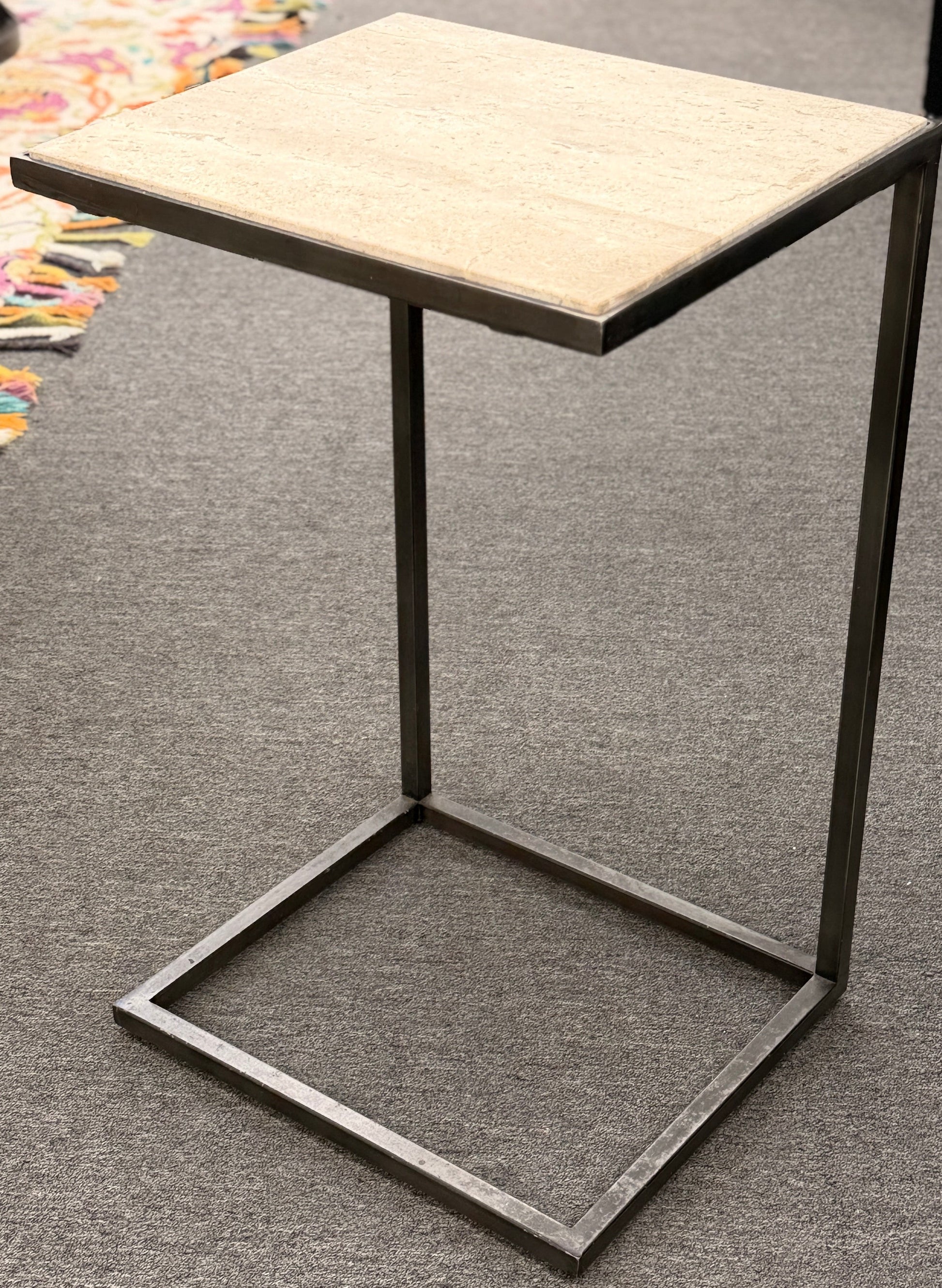 A contemporary C-shaped minimalist side table featuring a square tan stone top on a slender black metal frame designed to slide over a couch arm.
