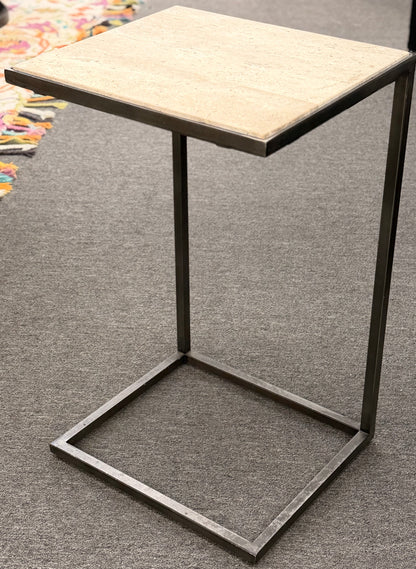 A contemporary C-shaped minimalist side table featuring a square tan stone top on a slender black metal frame designed to slide over a couch arm.