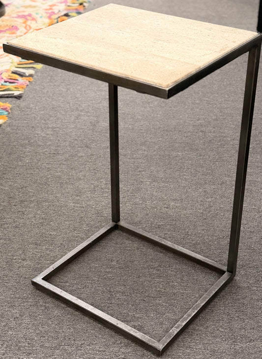 A contemporary C-shaped minimalist side table featuring a square tan stone top on a slender black metal frame designed to slide over a couch arm.