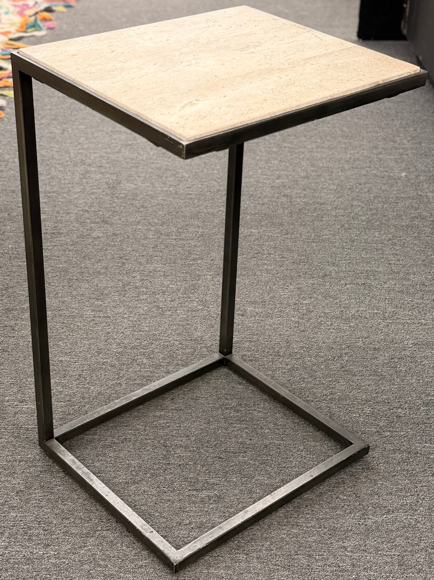A contemporary C-shaped minimalist side table featuring a square tan stone top on a slender black metal frame designed to slide over a couch arm.
