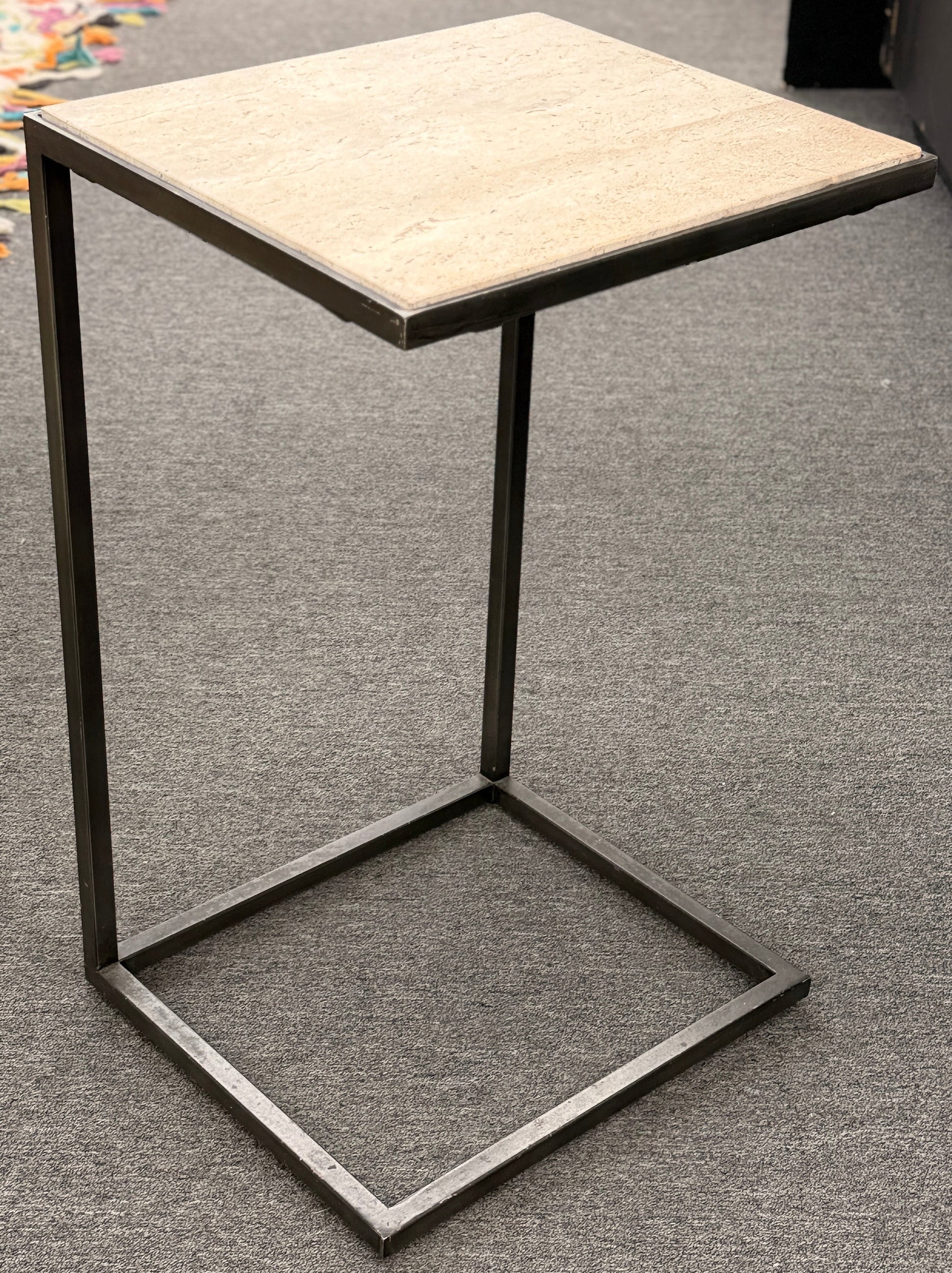 A contemporary C-shaped minimalist side table featuring a square tan stone top on a slender black metal frame designed to slide over a couch arm.