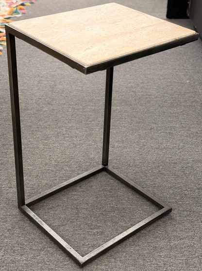 A contemporary C-shaped minimalist side table featuring a square tan stone top on a slender black metal frame designed to slide over a couch arm.