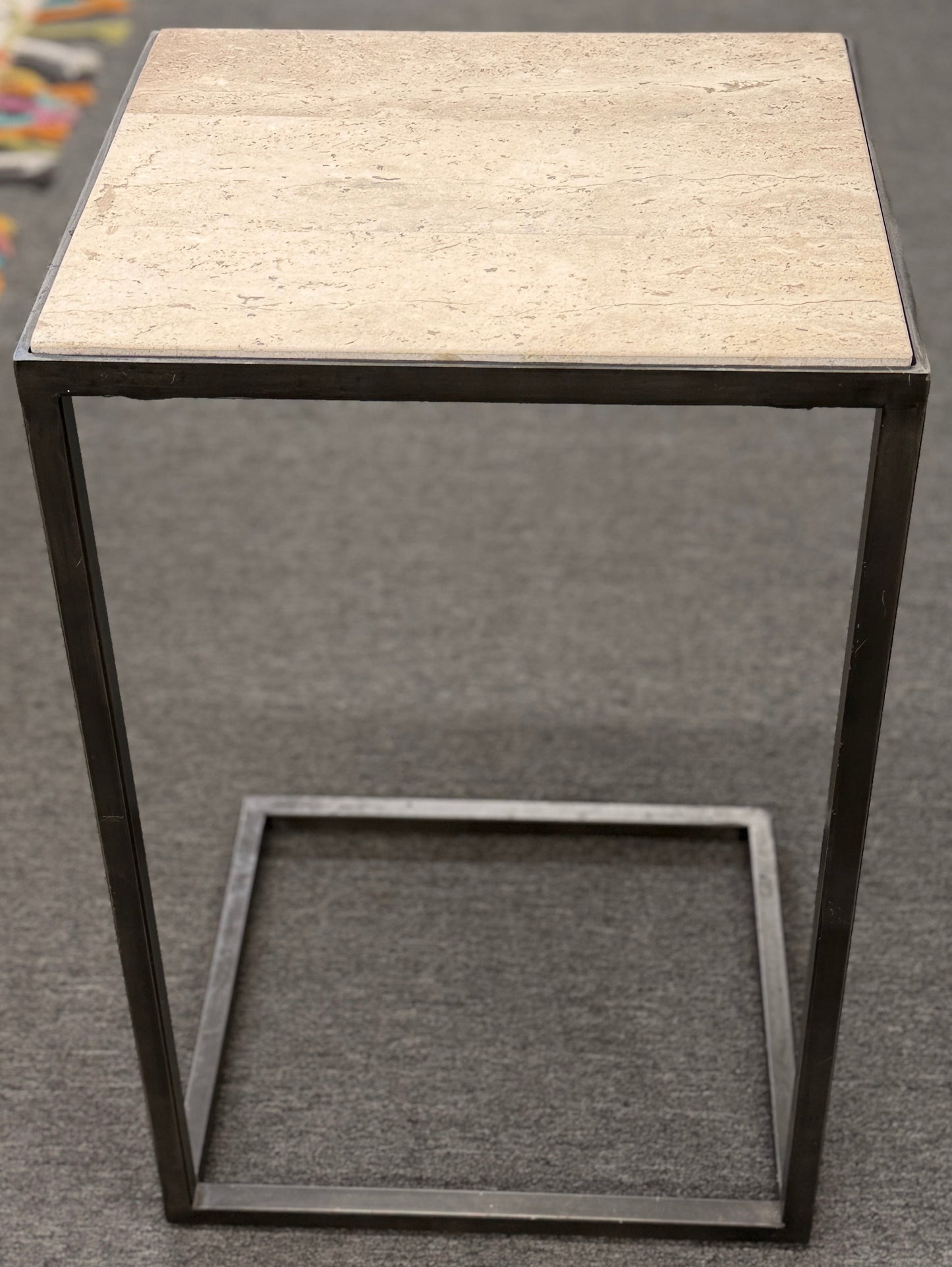 A contemporary C-shaped minimalist side table featuring a square tan stone top on a slender black metal frame designed to slide over a couch arm.