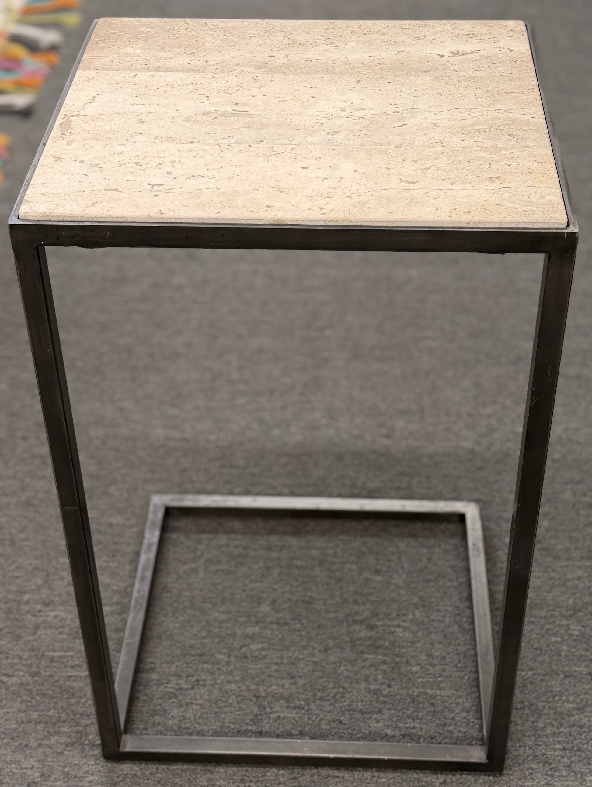 A contemporary C-shaped minimalist side table featuring a square tan stone top on a slender black metal frame designed to slide over a couch arm.