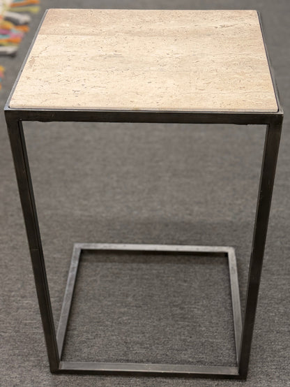 A contemporary C-shaped minimalist side table featuring a square tan stone top on a slender black metal frame designed to slide over a couch arm.