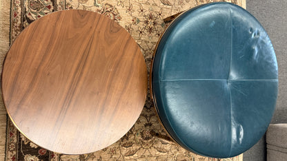 A two-piece contemporary nesting coffee table set featuring a petrol blue tufted leather round ottoman and a lower round walnut wood table on metal sled bases in good condition.
