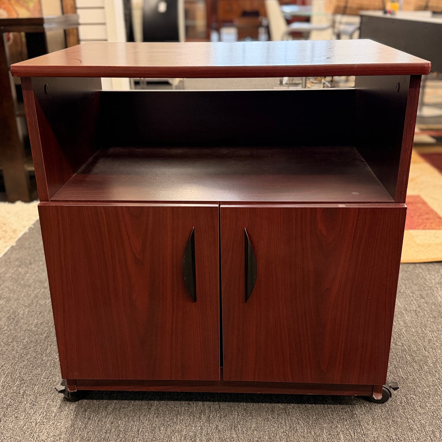 A compact mobile storage cabinet in a dark cherry finish featuring an open top shelf, double cabinet doors with curved black handles, and rolling wheels.