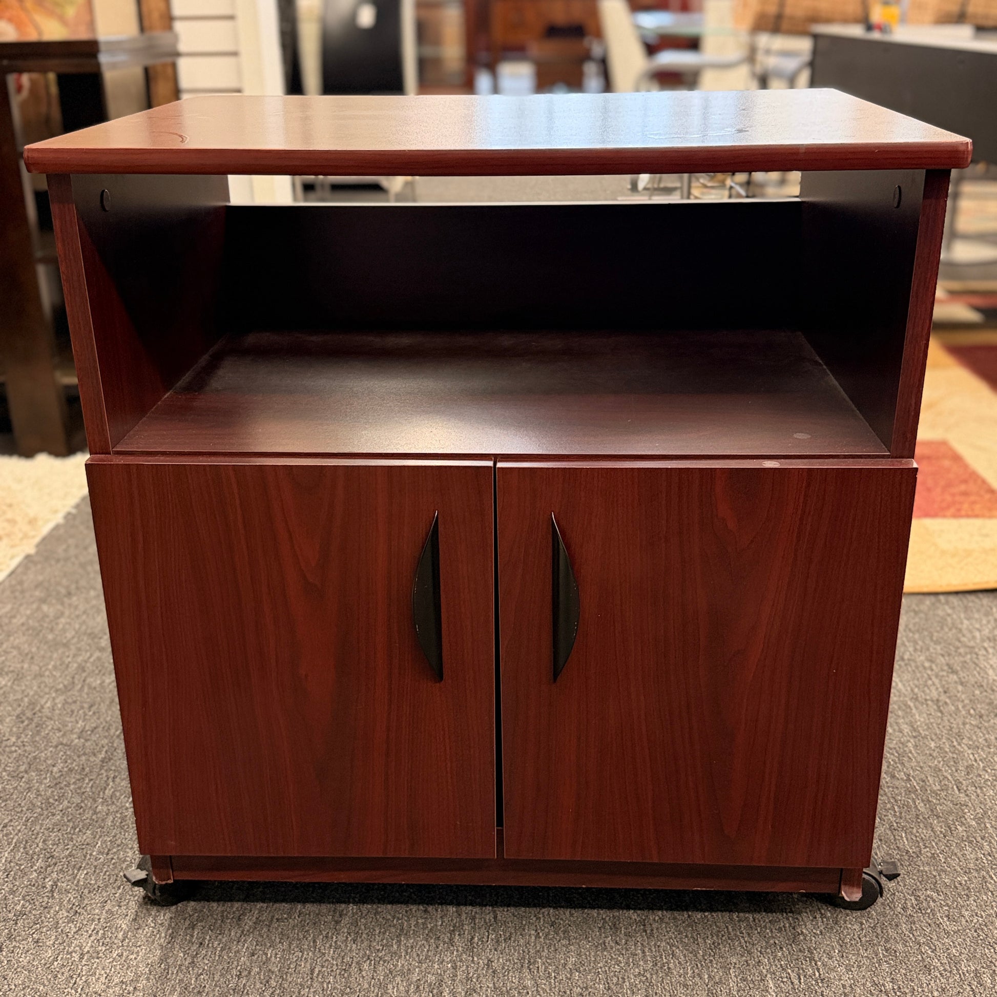 A compact mobile storage cabinet in a dark cherry finish featuring an open top shelf, double cabinet doors with curved black handles, and rolling wheels.