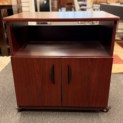 A compact mobile storage cabinet in a dark cherry finish featuring an open top shelf, double cabinet doors with curved black handles, and rolling wheels.