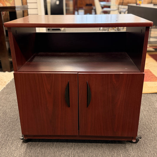 A compact mobile storage cabinet in a dark cherry finish featuring an open top shelf, double cabinet doors with curved black handles, and rolling wheels.