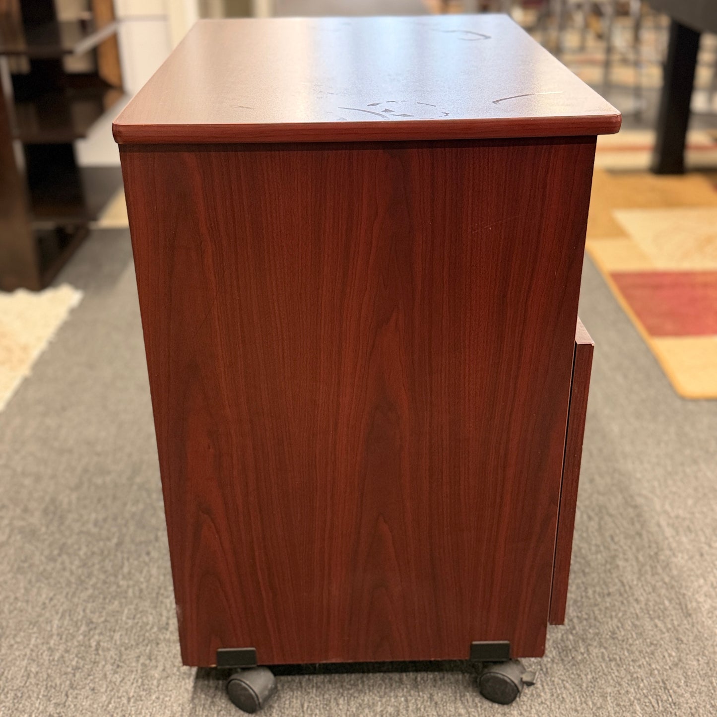 A compact mobile storage cabinet in a dark cherry finish featuring an open top shelf, double cabinet doors with curved black handles, and rolling wheels.