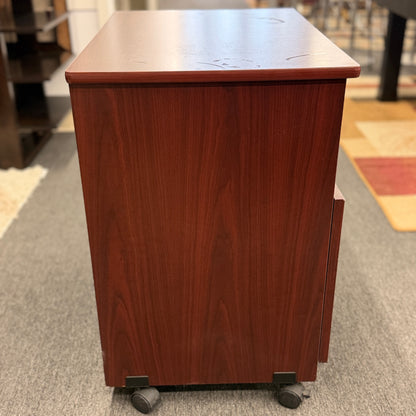 A compact mobile storage cabinet in a dark cherry finish featuring an open top shelf, double cabinet doors with curved black handles, and rolling wheels.