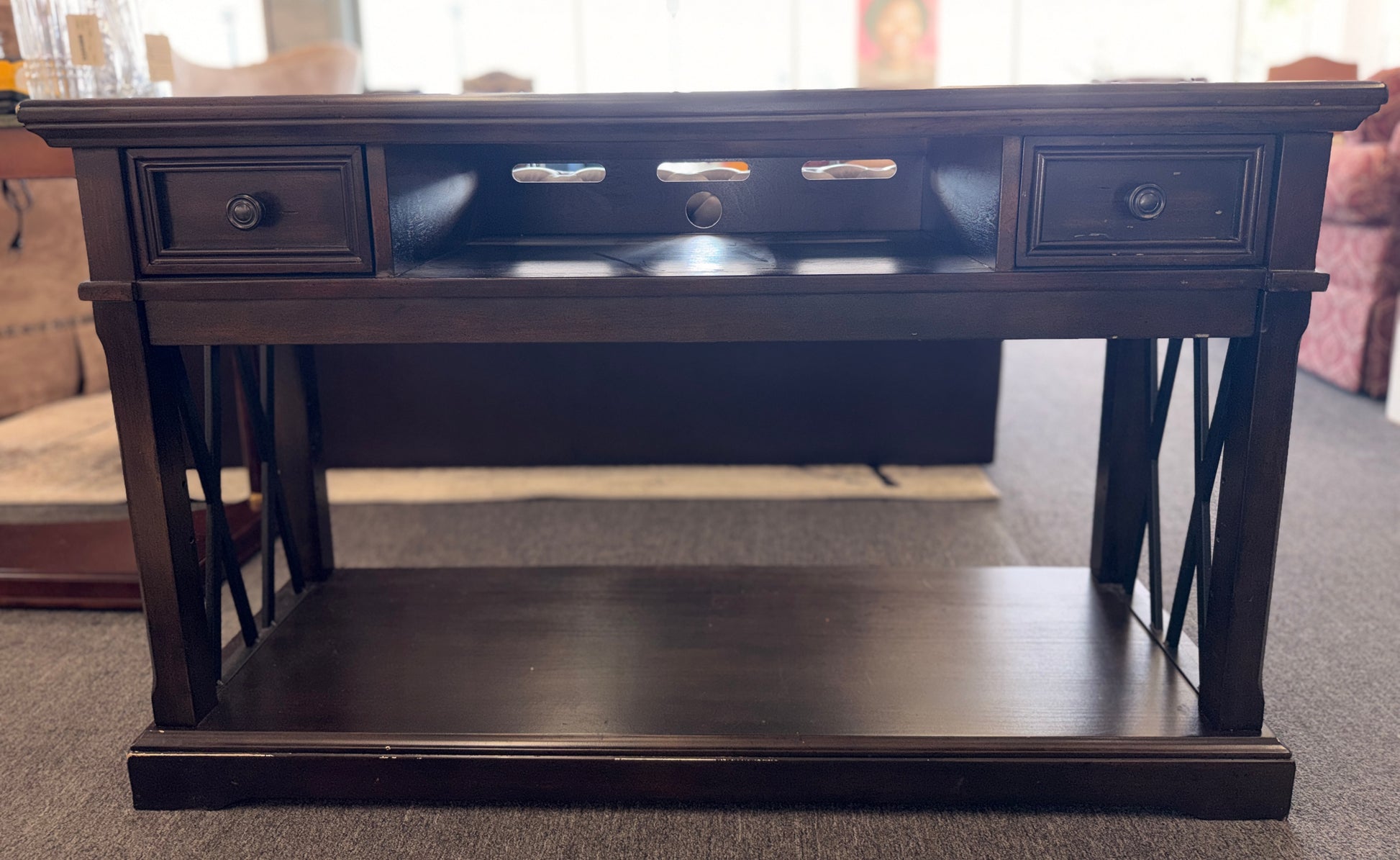 A dark-finished media console TV stand featuring X-brace side details, two small top drawers, a central open cubby with cable holes, and a large bottom shelf.