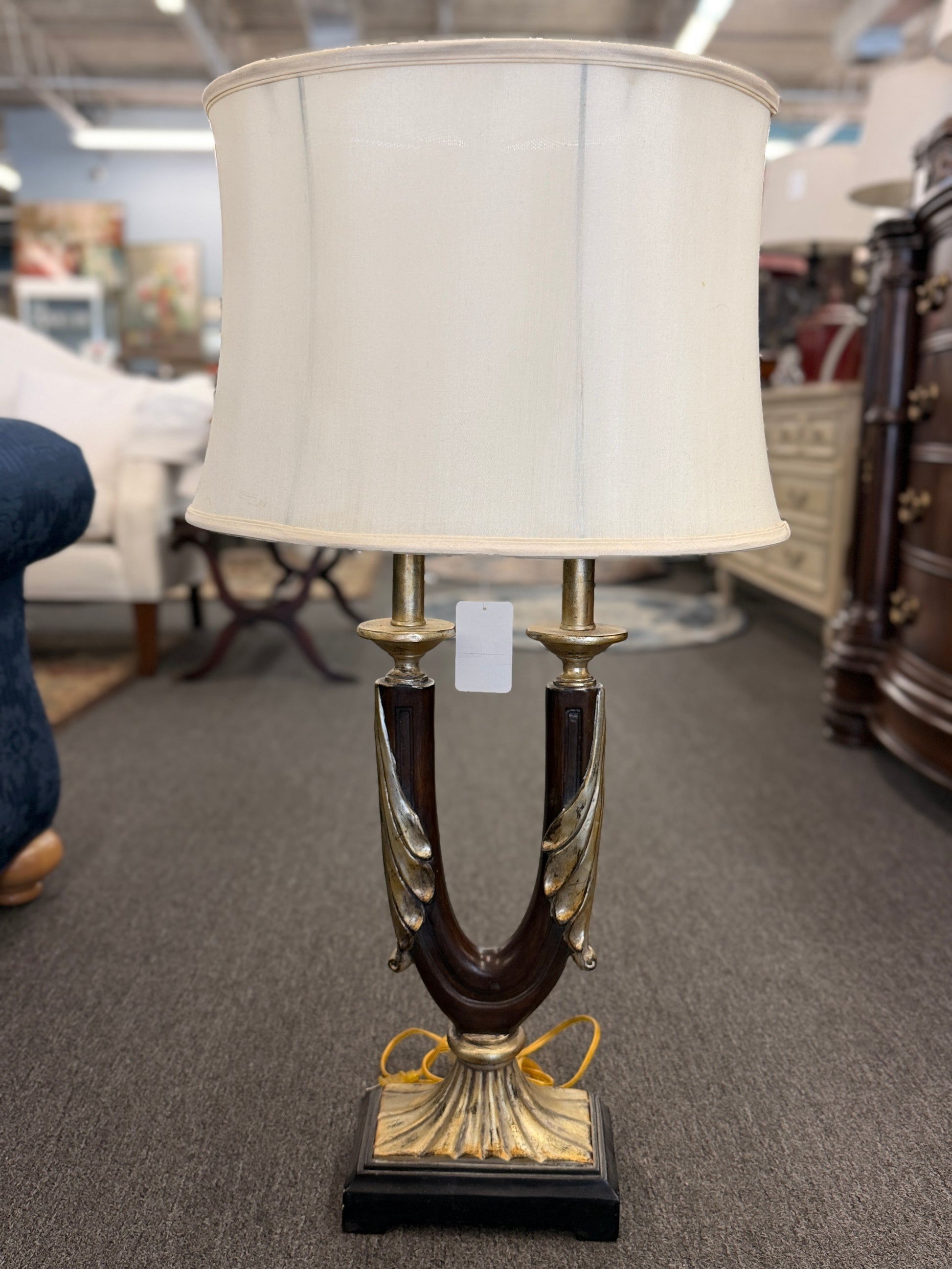 Decorative table lamp featuring a dark U-shaped body with metallic leaf accents, a fluted metallic pedestal, black square base, and a large cream-colored drum shade.